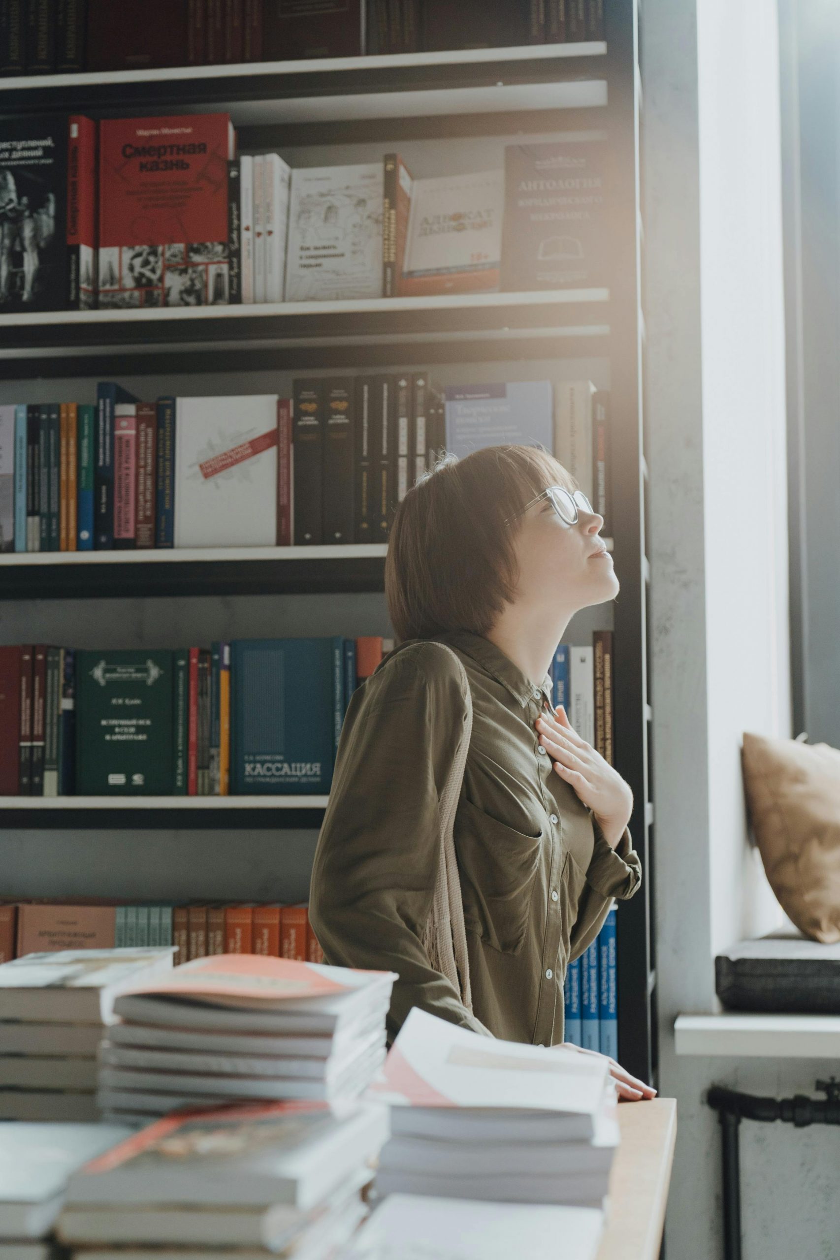 A woman stands thoughtfully in a bookstore, surrounded by books in a sunlit room.